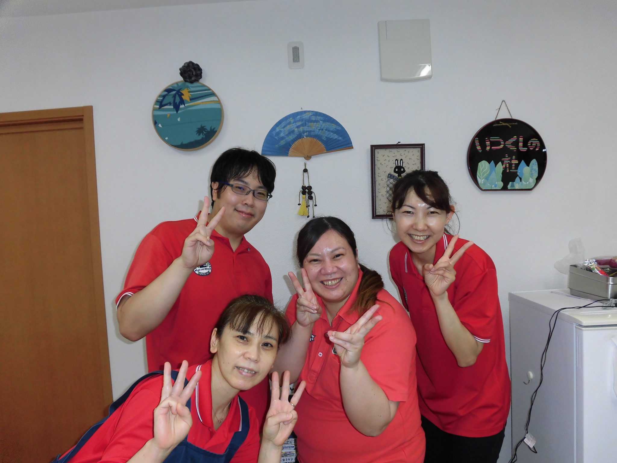 赤いユニフォームを着たスタッフ4名が笑顔でピースサインをしている。