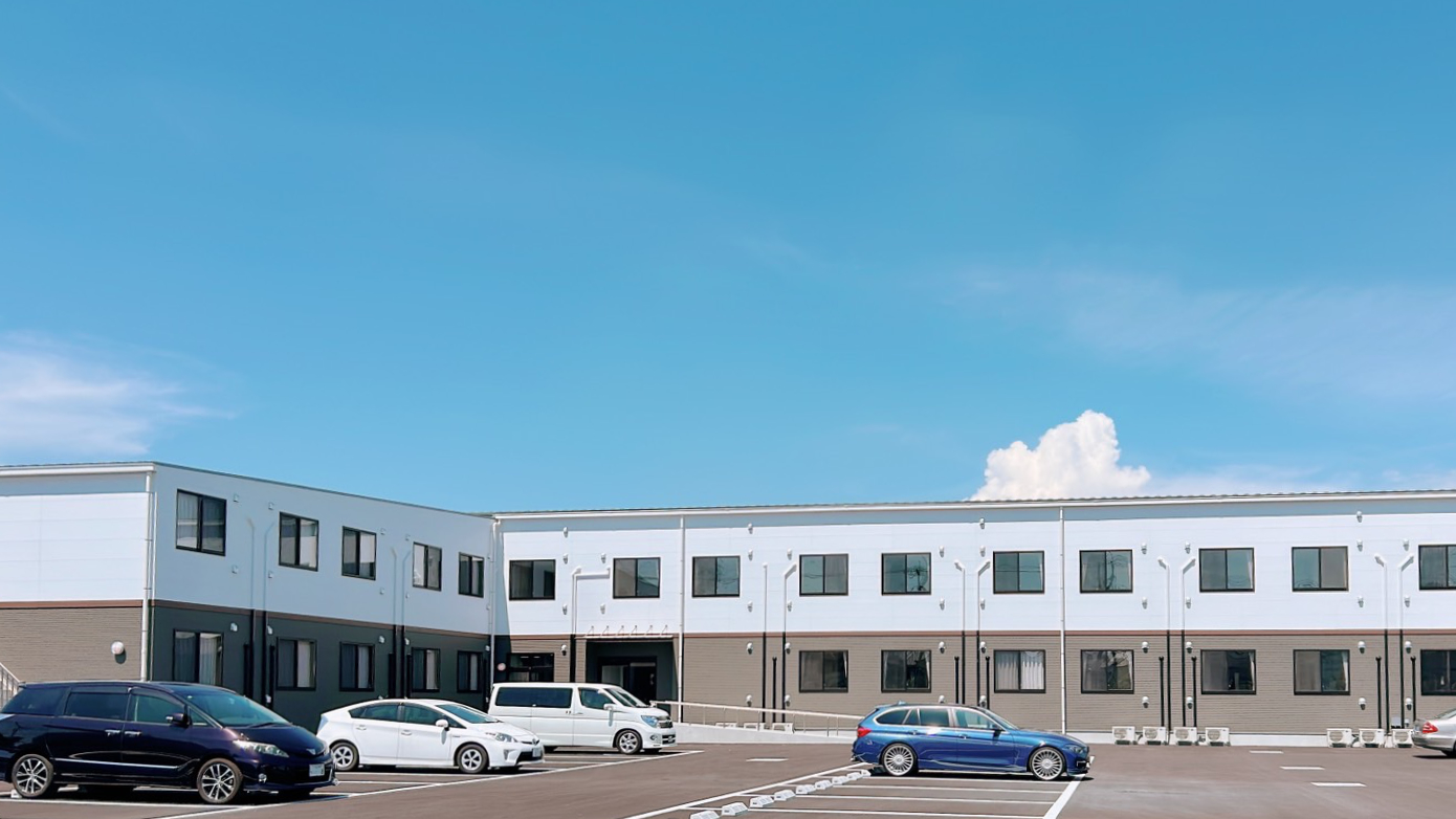 介護施設の建物外観。白とベージュ色の平屋建て、駐車場付き。